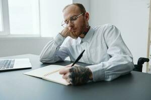 A man in a wheelchair, a businessman in the office works at a laptop, writing down a plan in a notebook, thoughtfulness, integration into society, the concept of working a person with disabilities photo