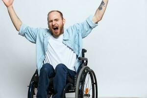 un hombre en un silla de ruedas mira a el cámara ira y agresión, con tatuajes en su brazos se sienta en un gris estudio fondo, salud concepto hombre con discapacidades foto