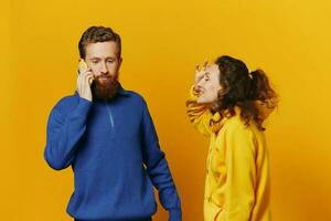 Man and woman couple with phone in hand call talking on the phone, on a yellow background, symbols signs and hand gestures, family quarrel jealousy and scandal. photo