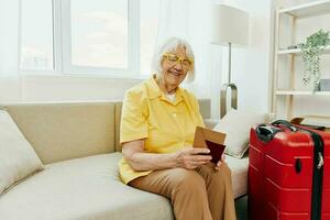 Happy senior woman with passport and travel ticket packed a red suitcase, vacation and health care. Smiling old woman joyfully sitting on the sofa before the trip raised her hands up in joy. photo