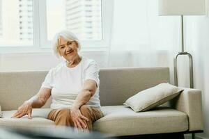 mayor mujer se sienta en un sofá a hogar en contra el fondo de un ventana y un felicidad sonrisa, elegante interior. estilo de vida Jubilación. foto