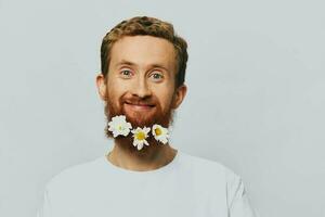 Portrait of a funny man in a white T-shirt with flowers daisies in his beard on a white isolated background, copy place. Holiday concept and congratulations. photo