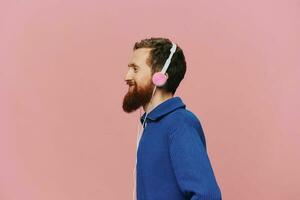 Portrait of a redheaded man wearing headphones smiling and dancing, listening to music on a pink background. A hipster with a beard. photo