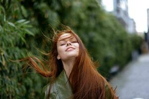 Portrait of a beautiful young girl in the city with a beautiful smile and red hair in a green raincoat in the city against a background of bamboo in spring, lifestyle in the city photo