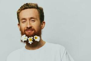 Portrait of a funny man in a white T-shirt with flowers daisies in his beard on a white isolated background, copy place. Holiday concept and congratulations. photo