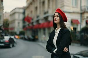 Moda mujer retrato en pie en el calle en frente de el ciudad en elegante ropa con rojo labios y rojo boina, viajar, cinematográfico color, retro Clásico estilo, urbano Moda estilo de vida. foto