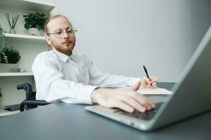 A man wheelchair businessman in the office works at a laptop, writing down ideas and a plan in a notebook, thoughtfulness, integration into society, the concept of working a person with disabilities photo