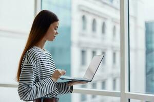 Woman with a laptop standing at the window in the office and looking out the window, work in an informal setting, business woman photo