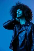 Portrait of fashion man with curly hair on blue background multinational, colored light, black leather jacket trend, modern concept. photo