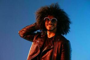 Portrait of a stylish man with curly hair with glasses smile on blue background multinational, color light, black leather jacket trend, modern concept. photo