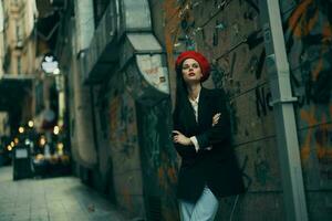 Moda mujer retrato caminando turista en elegante ropa con rojo labios caminando abajo un estrecho ciudad calle, viajar, cinematográfico color, retro Clásico estilo, dramático en contra un pared con pintada. foto