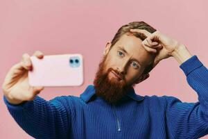 Portrait of a redheaded man with phone in hand taking selfies and photos on his phone with a smile on a pink background, blogger