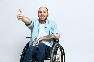 un hombre en un silla de ruedas mira a el cámara muestra un pulgares arriba, felicidad, con tatuajes en su manos se sienta en un gris estudio fondo, salud concepto hombre con discapacidades foto