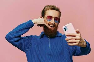 Portrait of a redheaded man with phone in hand taking selfies and photos on his phone with a smile on a pink background, blogger