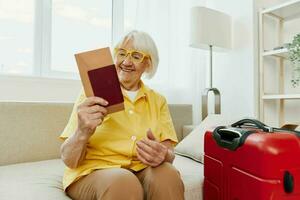 Happy senior woman with passport and travel ticket packed a red suitcase, vacation and health care. Smiling old woman joyfully sitting on the sofa before the trip raised her hands up in joy. photo