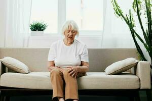 mayor mujer grave dolor en su pierna sentado en el sofá, salud problemas en antiguo edad, pobre calidad de vida. abuela con gris pelo sostiene en a su dolorido rodilla, problemas con articulaciones y ligamentos foto