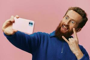 Portrait of a redheaded man with phone in hand taking selfies and photos on his phone with a smile on a pink background, blogger