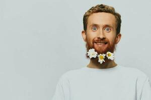 Portrait of a funny man in a white T-shirt with flowers daisies in his beard on a white isolated background, copy place. Holiday concept and congratulations. photo