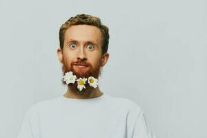 Portrait of a funny man in a white T-shirt with flowers daisies in his beard on a white isolated background, copy place. Holiday concept and congratulations. photo