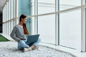 A woman works online via the Internet on her laptop and smiles looking out the large windows of a tall building, an urban style concept and working in an office photo
