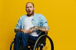 un hombre en un silla de ruedas sorprendido, con tatuajes en su brazos se sienta en un amarillo estudio fondo, el concepto de salud es un persona con discapacidades foto