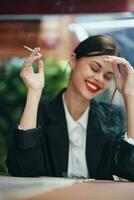 elegante mujer se sienta en un café a un mesa y fuma un cigarrillo liberando fumar desde su boca, un malo hábito, un sonrisa con dientes y un pensativo Mira foto