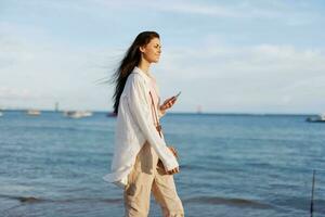 Lanza libre mujer con teléfono en mano en vacaciones caminando en el playa por el Oceano en bali, contento viaje y vacaciones, móvil comunicación foto