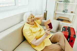 contento mayor mujer con pasaporte y viaje boleto lleno un rojo maleta, vacaciones y salud cuidado. sonriente antiguo mujer alegremente sentado en el sofá antes de el viaje elevado su manos arriba en alegría. foto