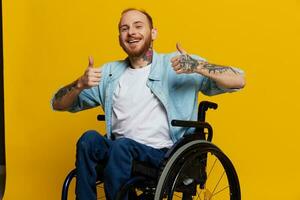 un hombre en un silla de ruedas sonrisa y felicidad, pulgar arriba, con tatuajes en su manos se sienta en un amarillo estudio fondo, el concepto de salud un persona con discapacidades foto