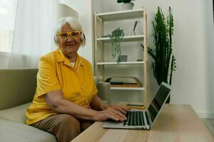 Happy elderly woman with a laptop typing with headphones sitting at home on the couch in a yellow shirt, bright modern interior, lifestyle online communication. photo