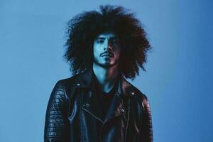 Portrait of fashion man with curly hair on blue background multinational, colored light, black leather jacket trend, modern concept. photo