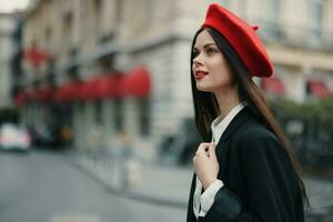 Moda mujer retrato belleza en pie en el calle en frente de el ciudad en elegante ropa con rojo labios y rojo boina, viajar, cinematográfico color, retro Clásico estilo, urbano Moda estilo de vida. foto