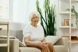 mayor mujer grave dolor en su pierna sentado en el sofá, salud problemas en antiguo edad, pobre calidad de vida. abuela con gris pelo sostiene en a su dolorido rodilla, problemas con articulaciones y ligamentos foto