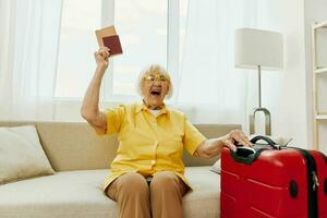 Happy senior woman with passport and travel ticket packed a red suitcase, vacation and health care. Smiling old woman joyfully sitting on the sofa before the trip raised her hands up in joy. photo