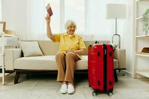 elderly woman with passport and travel ticket packed a red suitcase, vacation and health care. Smiling old woman joyfully sitting on the sofa before the trip raised her hands up the joy of retirement. photo