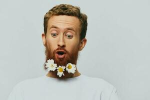 Portrait of a funny man in a white T-shirt with flowers daisies in his beard on a white isolated background, copy place. Holiday concept and congratulations. photo