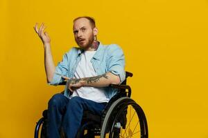 A man in a wheelchair problems with the musculoskeletal system looks at the camera is not satisfied, with tattoos on his arms sits on a yellow studio background, health concept man with disabilities photo
