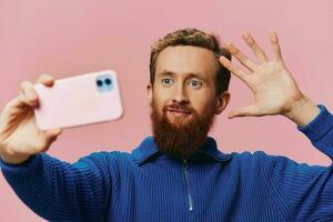 Portrait of a redheaded man with phone in hand taking selfies and photos on his phone with a smile on a pink background, blogger