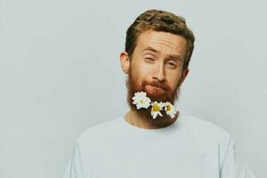 Portrait of a funny man in a white T-shirt with flowers daisies in his beard on a white isolated background, copy place. Holiday concept and congratulations. photo