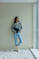 Freelance woman holding a laptop standing full-length at a large window and looking out at tall buildings in the city, work and study online photo