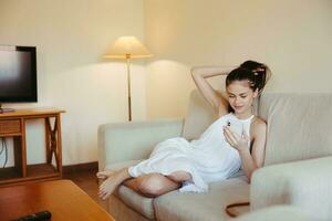 A young woman sits on the couch in the living room with her phone in her hands, relaxing online work and social media freelance blogger. photo