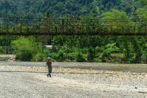 Man standing near the river with old hanging bridge, in Lokop Serbajadi, east Aceh, Indonesia photo