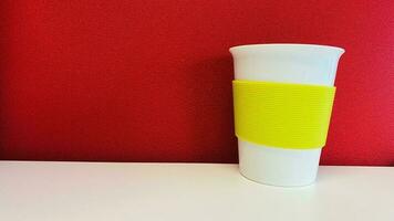 A white paper cup with a yellow border on a white table. Background red wall. photo