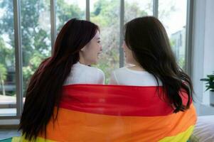 el lgbt Pareja se sentó en el cama, cubierto en arco iris banderas, mirando fuera el ventana a observar el naturaleza en el hotel habitación. foto