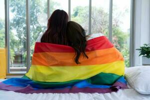 el lgbt Pareja se sentó en el cama, cubierto en arco iris banderas, mirando fuera el ventana a observar el naturaleza en el hotel habitación. foto