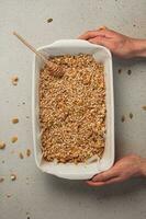 Hands holding a baking dish with oatmeal, cashews, pumpkin seeds, raisins and a honey spoon on a white surface. Ingredients for homemade granola. Top view. Perfect for stories and vertical wallpaper photo