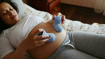 parte superior ver de un embarazada mujer poniendo bebé botines en su barriga en tarde el embarazo tercero trimestre, disfrutando su maternidad estilo de vida, relajante a hogar, acostado en el cama a hogar dormitorio.esperando un bebé video