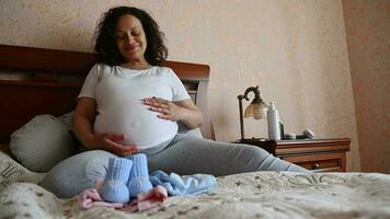 Happy delightful multi ethnic woman caressing her belly, relaxing sitting on the bed in a tranquil homely atmosphere, enjoying maternity lifestyle. Pregnancy trimester third. Expectation of a baby video