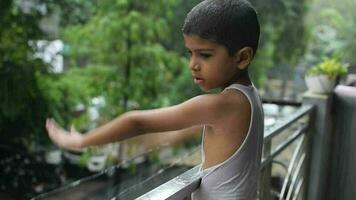 pequeño niño jugando en verano lluvia en casa balcón, indio inteligente chico jugando con lluvia gotas durante monzón lluvioso estación, niño jugando en lluvia video