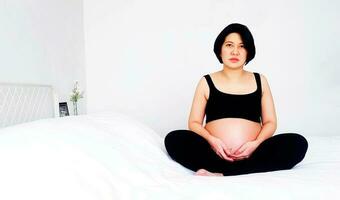 asiático embarazada mujer en negro camiseta sentado en blanco cama y conmovedor su barriga en dormitorio con Copiar espacio. el embarazo 7-8 meses, maternidad, amar, expectativa, cuidado y esperando para su niño. foto